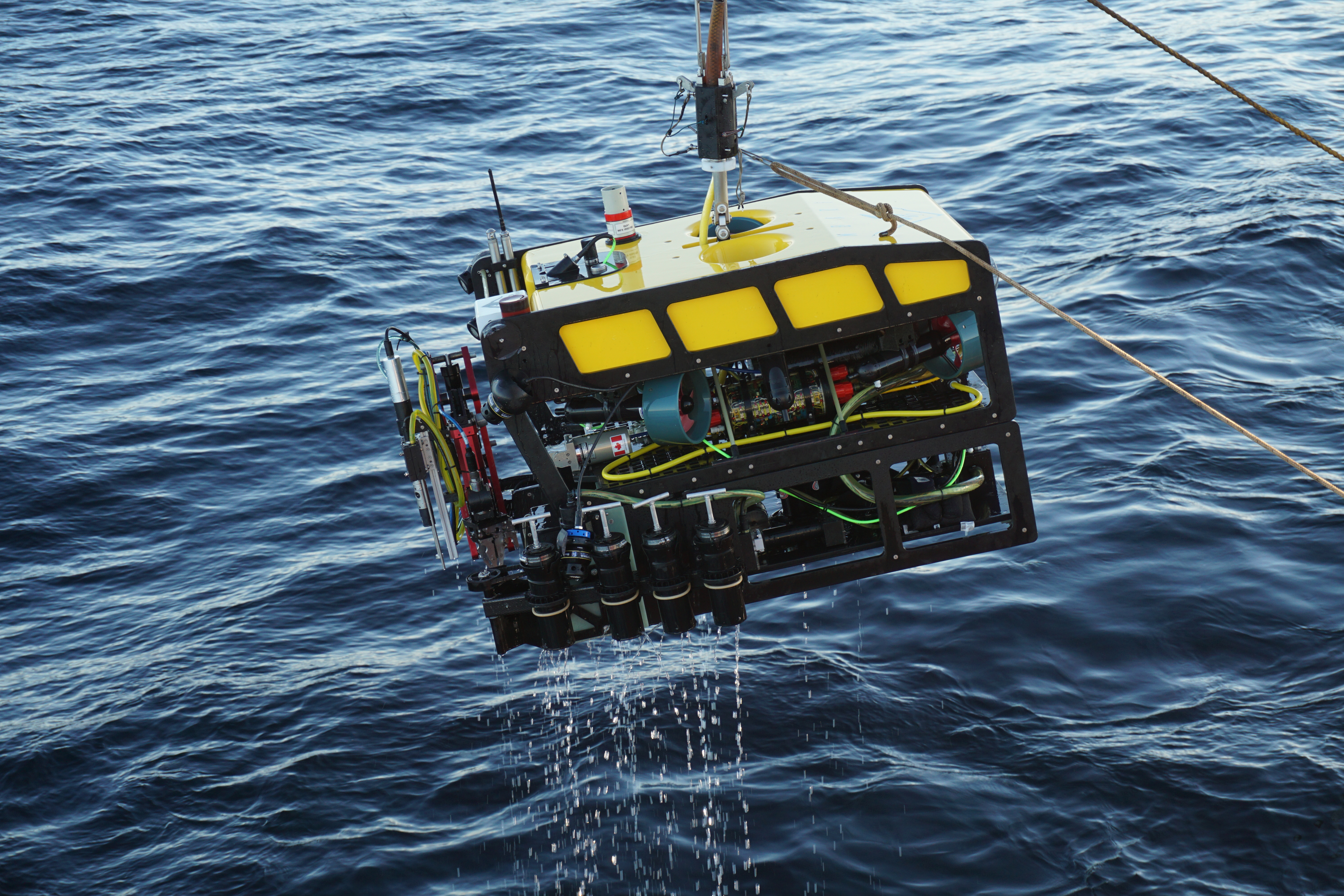 A yellow and black remotely operate vehicle is suspended over the water.