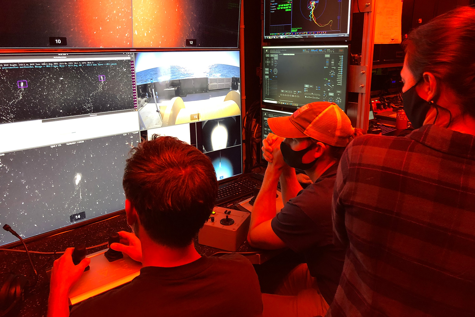 Three people wearing face black surgical face masks star at a series of screens in a room with red lighting.