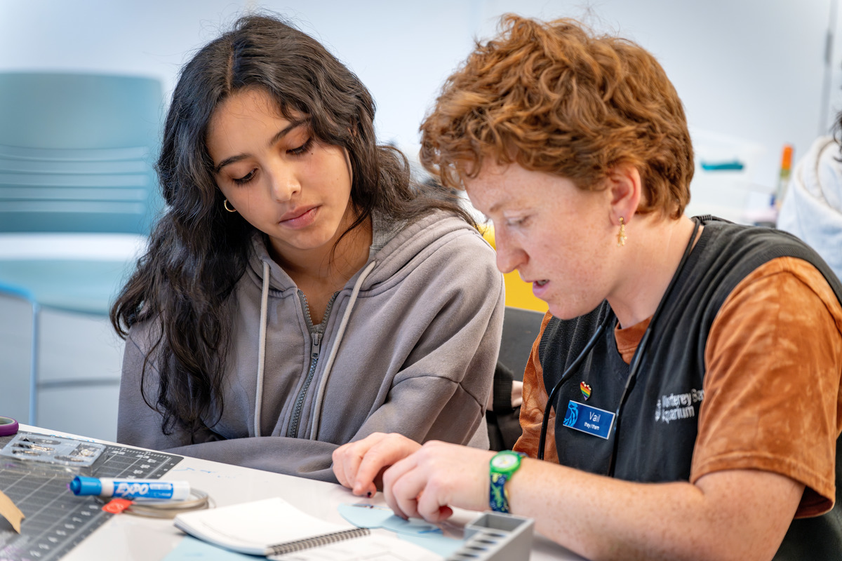 A blue-uniformed Aquarium staff member and a teen lean over a table working on a project together
