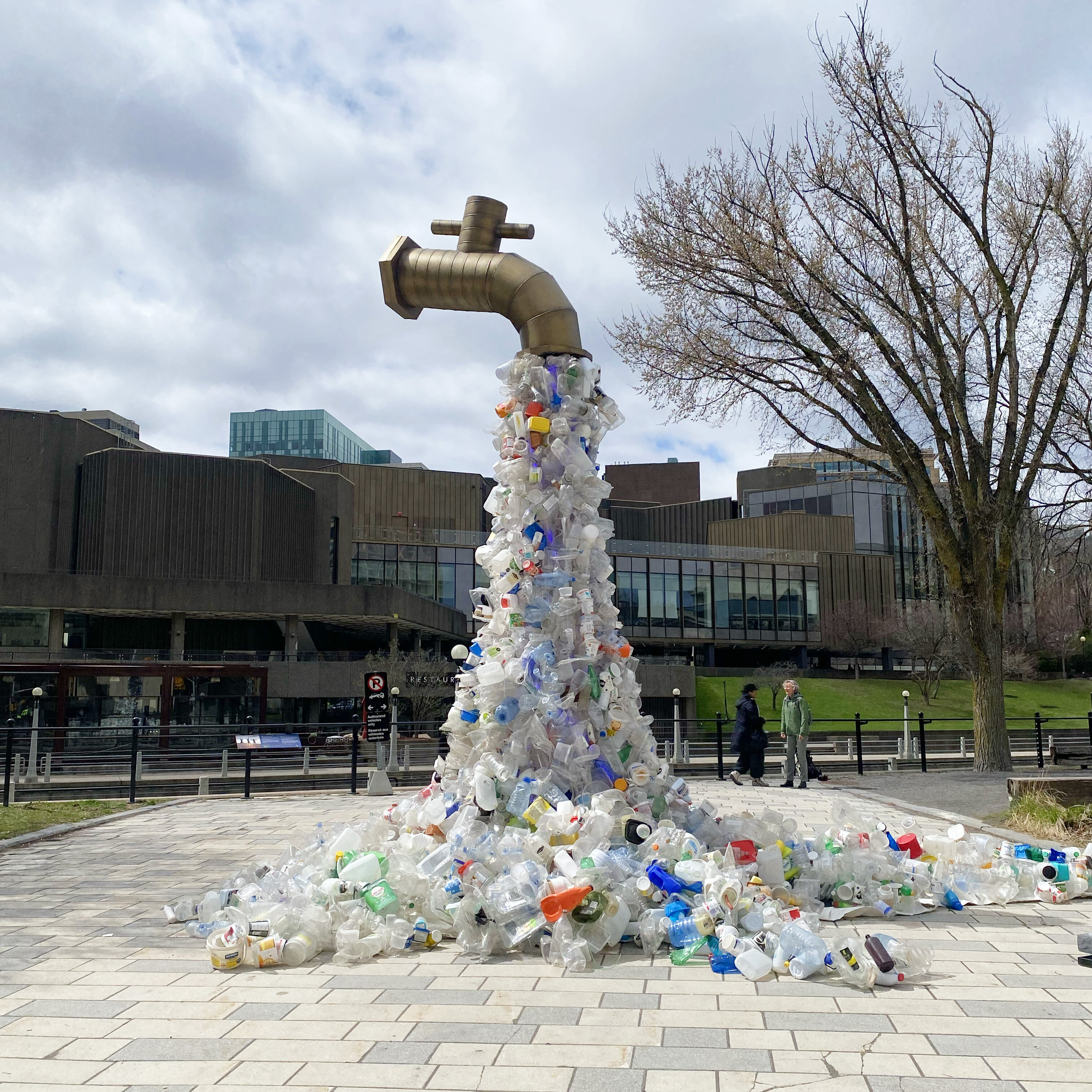 Large outdoor sculpture of a giant faucet pouring a cascade of plastic bottles and containers onto the ground