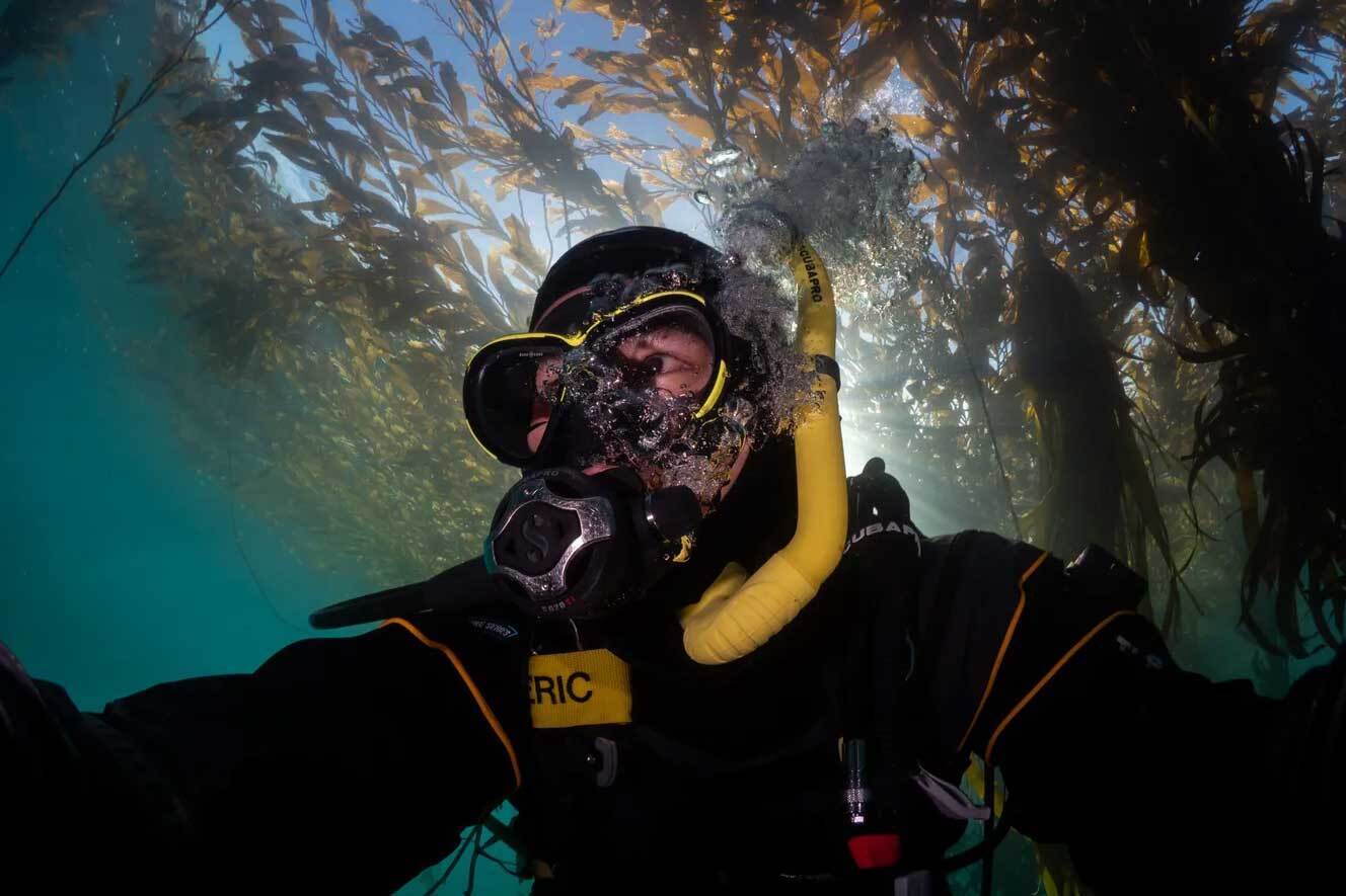 Underwater selfie of Eric swimming in Monterey Bay Kelp Forest with full diving gear and sunlight filters through swaying kelp behind