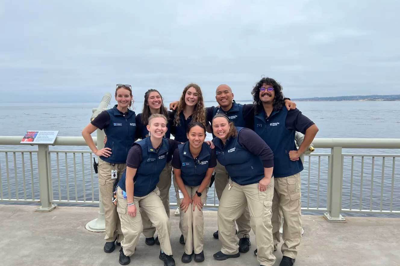 2022 Aquarium Naturalist cohort poses in two rows, arms linked, on the back deck with ocean view behind