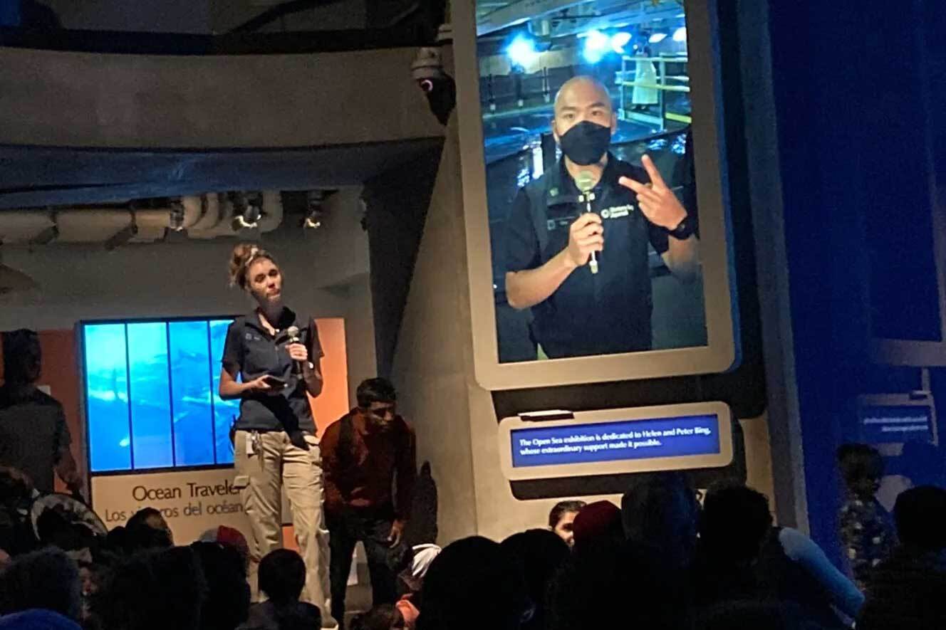 Eric flashes a peace sign with one hand and holds a microphone in the other as the audience watches on a vertical screen by the Open Sea while colleague stands nearby with a microphone and phone