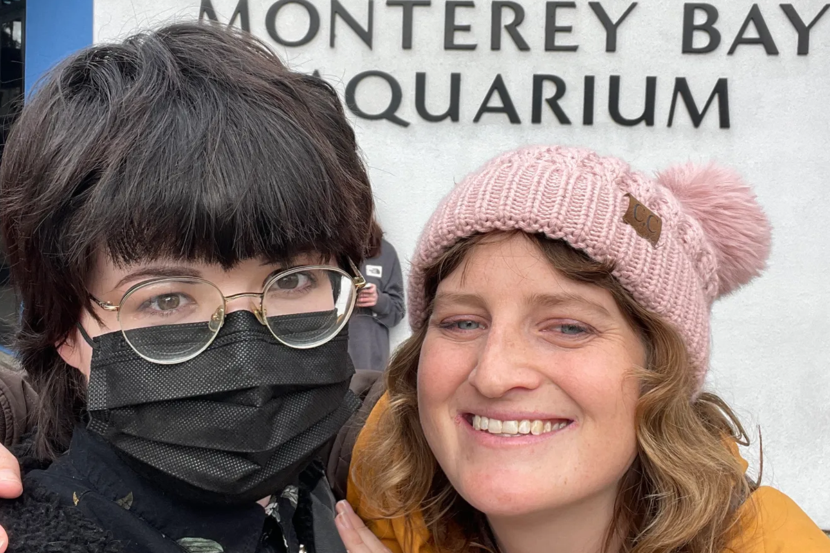 Lilianna and her girlfriend, Allison, take a selfie in front of the Aquarium entrance. The Aquarium's logo and name is visible behind the couple.