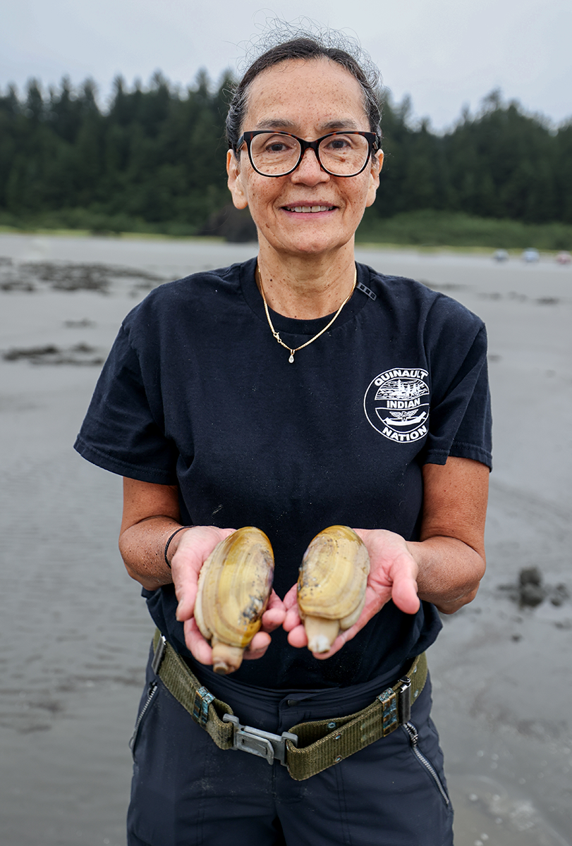 A smiling woman in a Quinault Indian Nation shirt holds two razor clams toward the camera on a beach