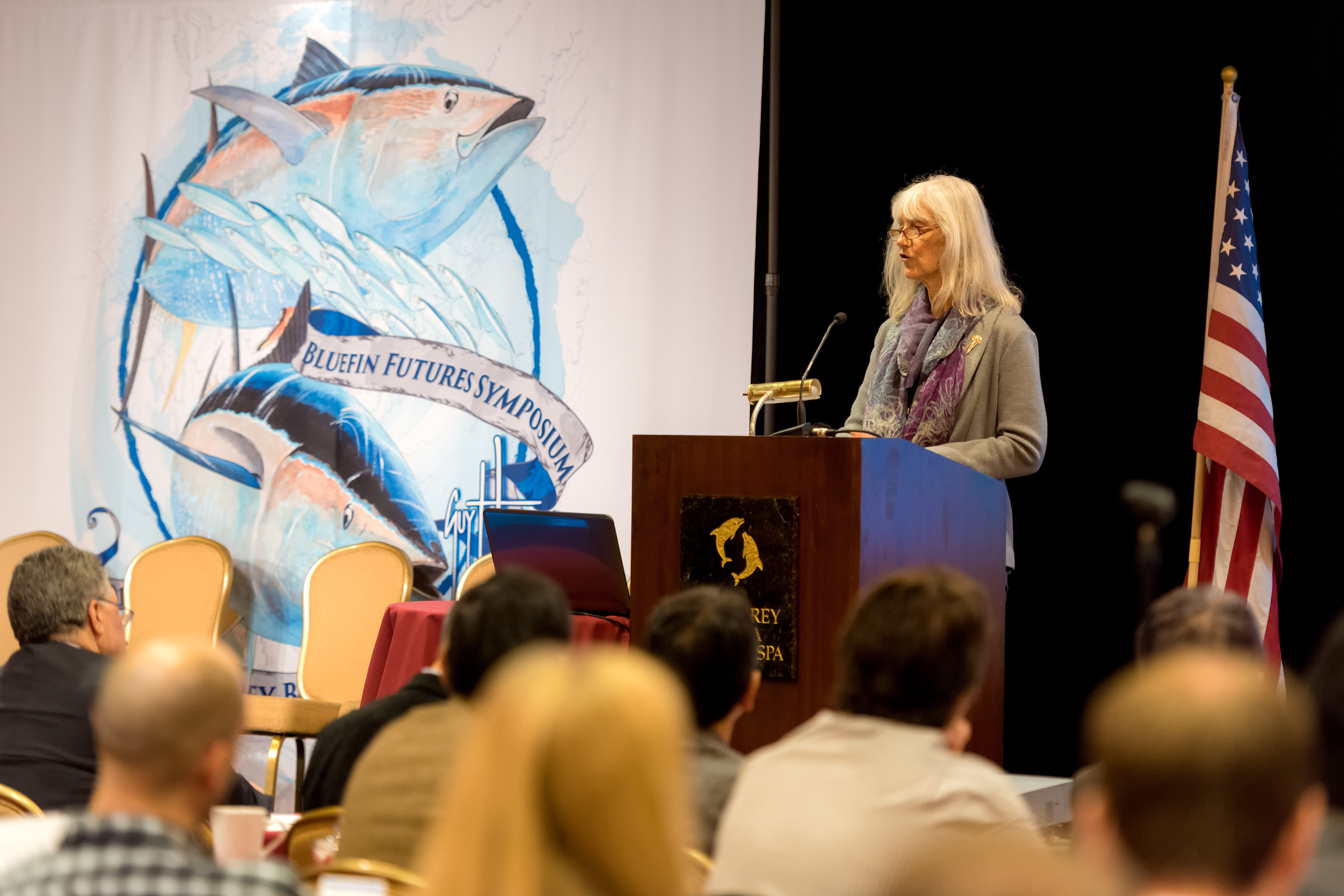 Julie Packard speaks at a podium during the Bluefin Futures Symposium, with an illustrated banner of bluefin tuna and a US flag in the background and audience members are seated and listening