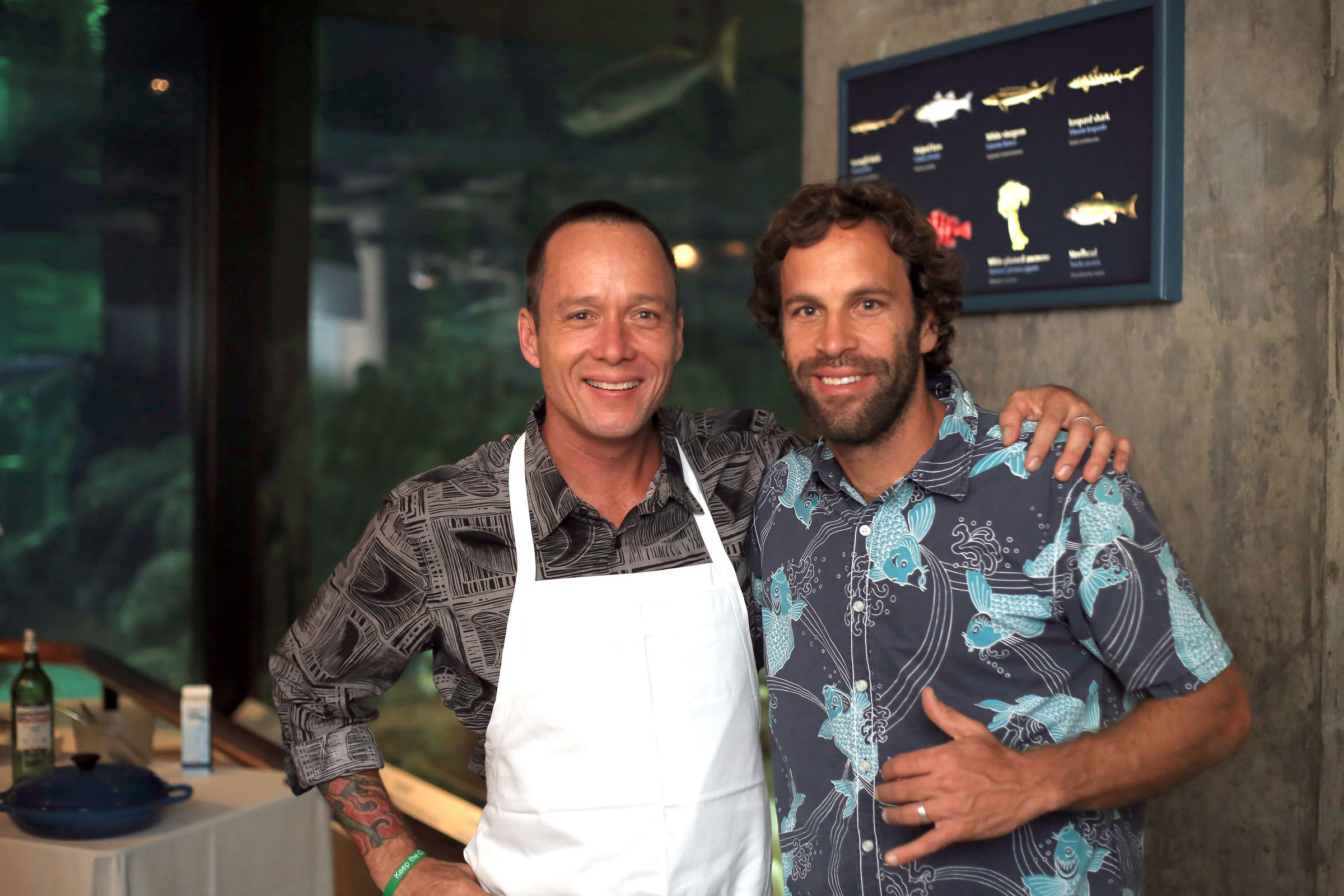 Blue Ribbon Task Force chef Ed Kenney and singer Jack Johnson standing side by side indoors. One wears an apron. Behind them is an aquarium and a wall display with fish illustrations