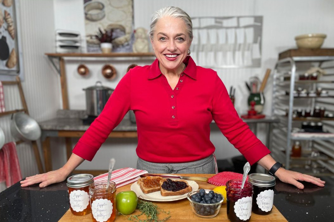 Blue Ribbon Task Force chef Virginia Willis in a red shirt stands in a kitchen with a wooden board displaying jars of jams, toast, an apple, blueberries, and herbs
