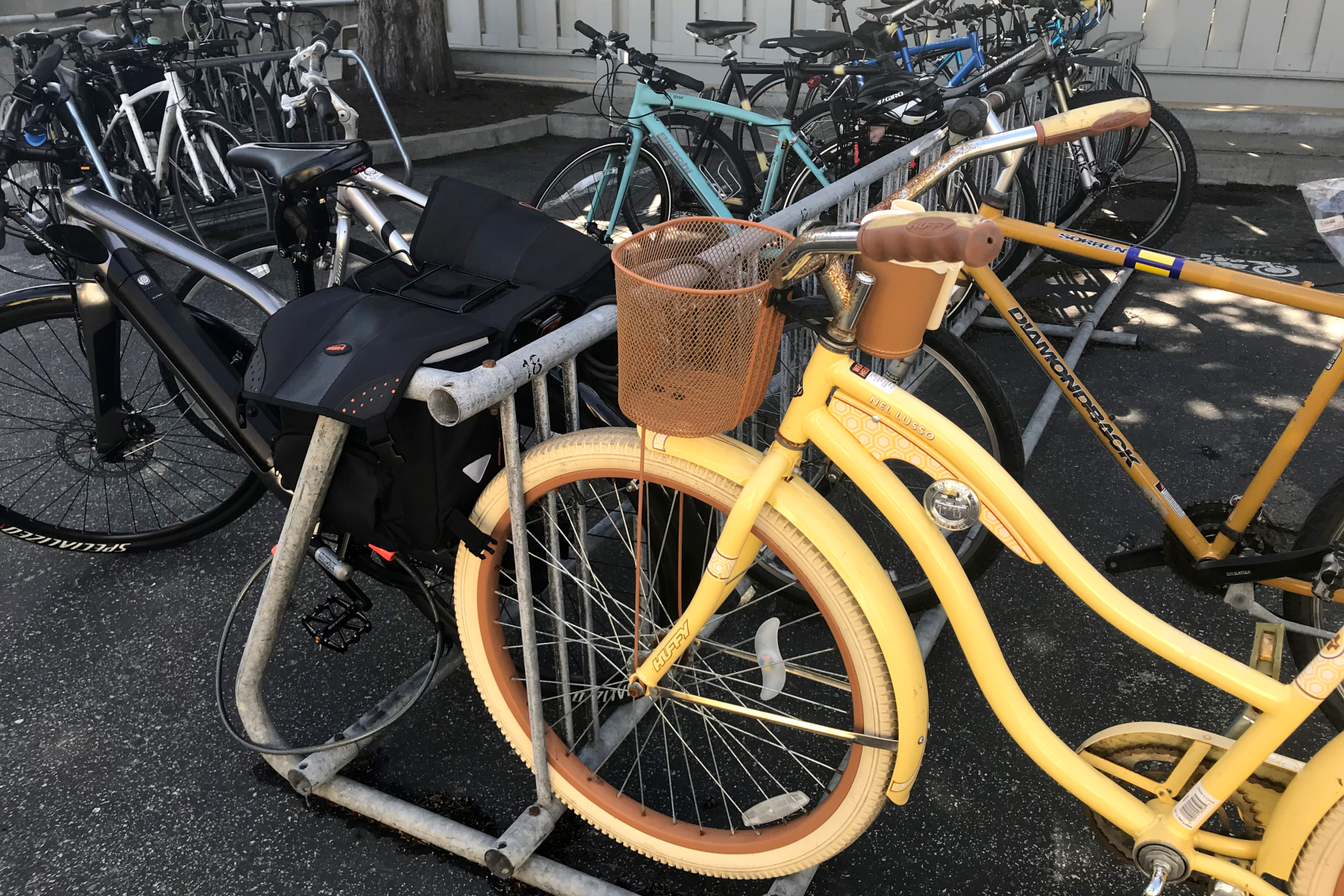 A yellow cruiser bike with tan tires and a mesh front basket locked to a metal rack, surrounded by other bicycles