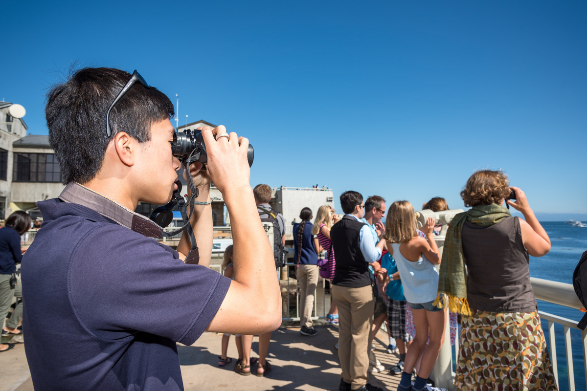 A young man looks through binoculars on a sunny outdoor deck and in the background, a crowd of people gathers along the railing to view the ocean