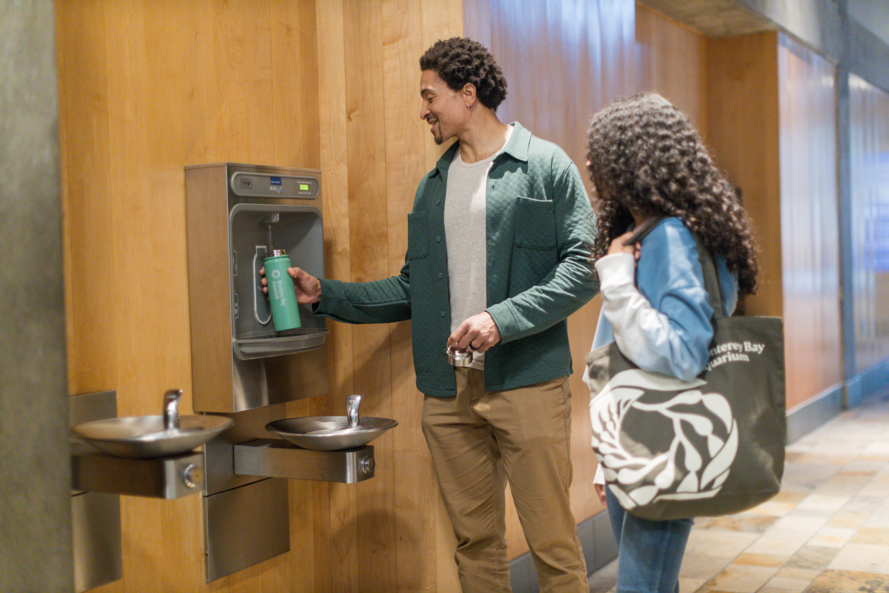 A visitor refilling their water bottle while another visitor stands close by waiting holding an Aquarium tote bag
